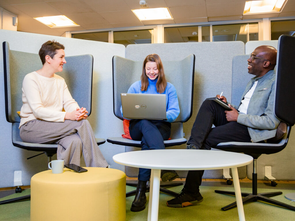 Three people sitting on chairs chatting, the middle one looking at her computer.