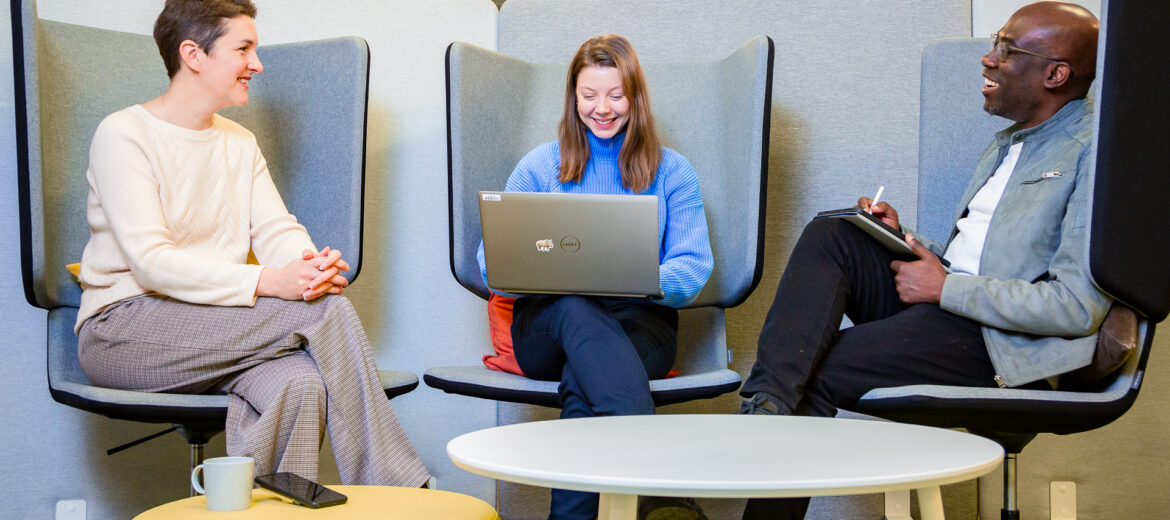 Three people sitting on chairs chatting, the middle one looking at her computer.