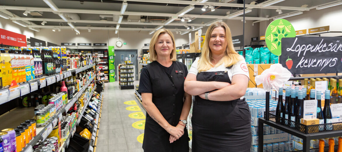 Alina ja Anna seicovat Alkon hyllyriven välissä ja hymyilevät kameralle.