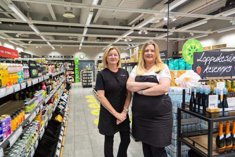 Alina ja Anna seicovat Alkon hyllyriven välissä ja hymyilevät kameralle.